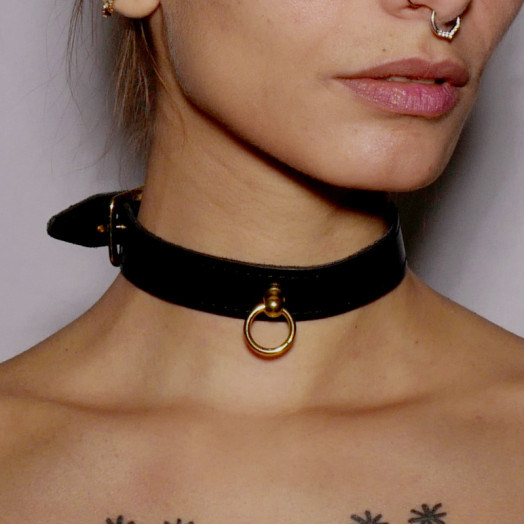 Choker fin en cuir noir surmonté d'un anneau en acier doré. Il se porte comme bijou ou accessoire de jeu de soumission